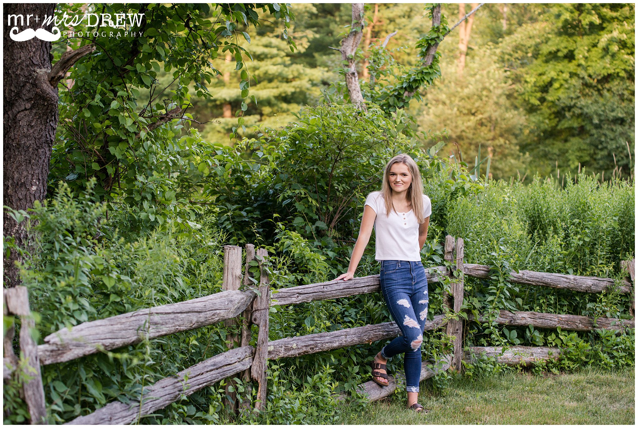 Senior Portrait of girl in white t-shirt and ripped jeans leaning on wooden fence. 
