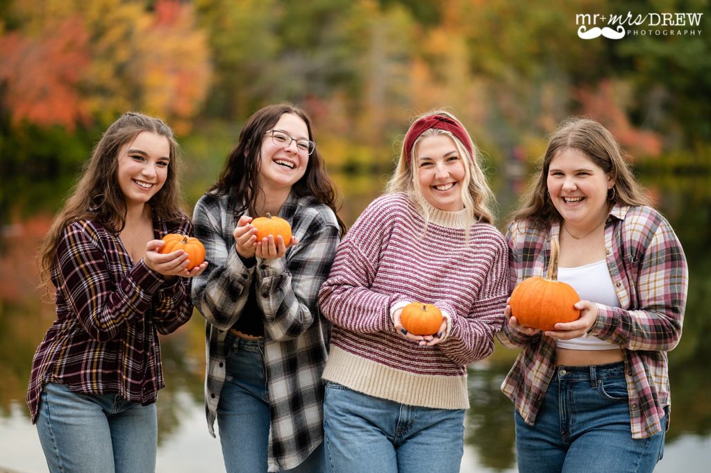 senior girls holding tiny pumpkins and smiling at Heart Pond Chelmsford MA