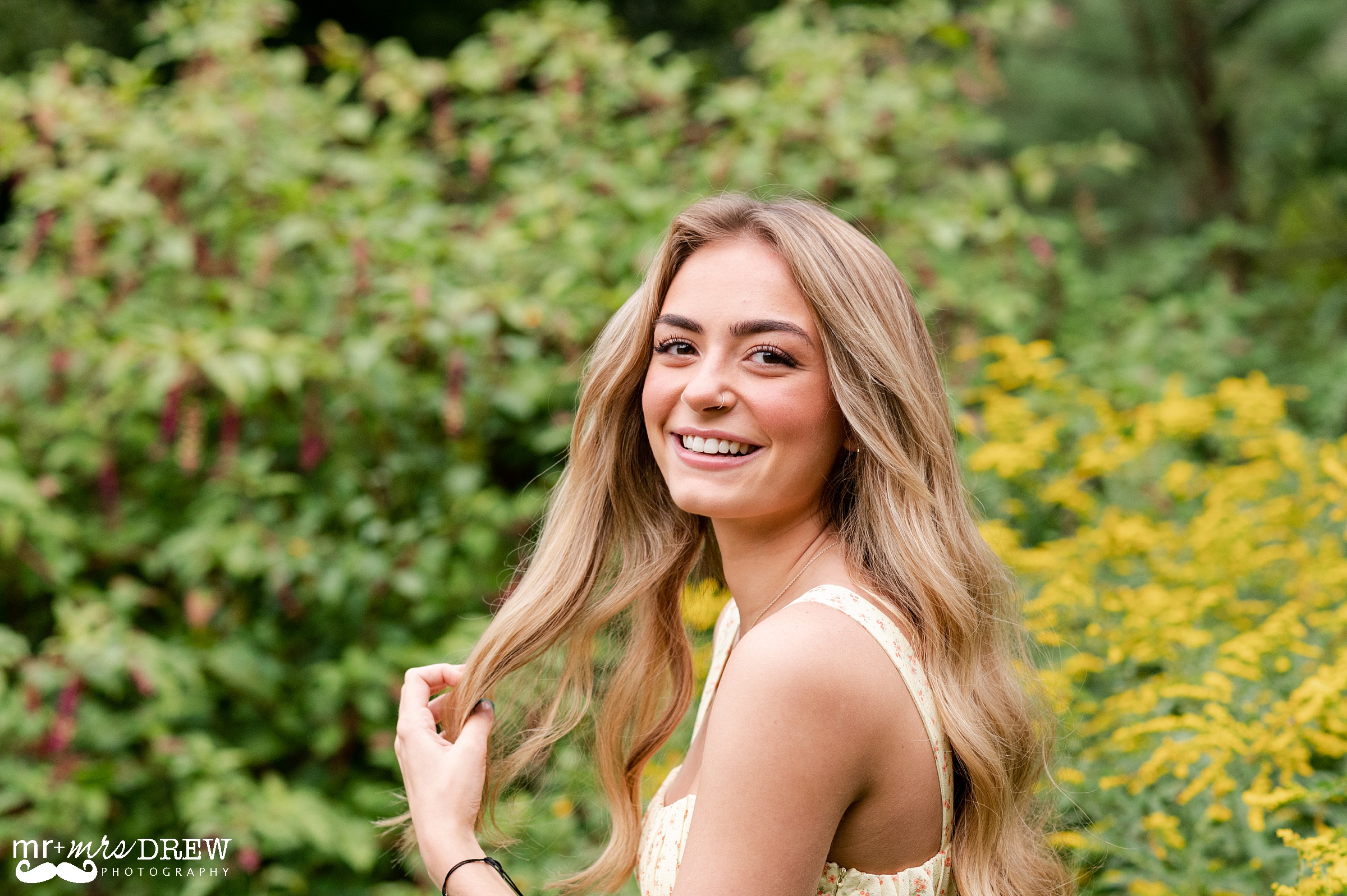 Senior twirling her hair while smiling at camera in a yellow dress with a yellow dress in background. 