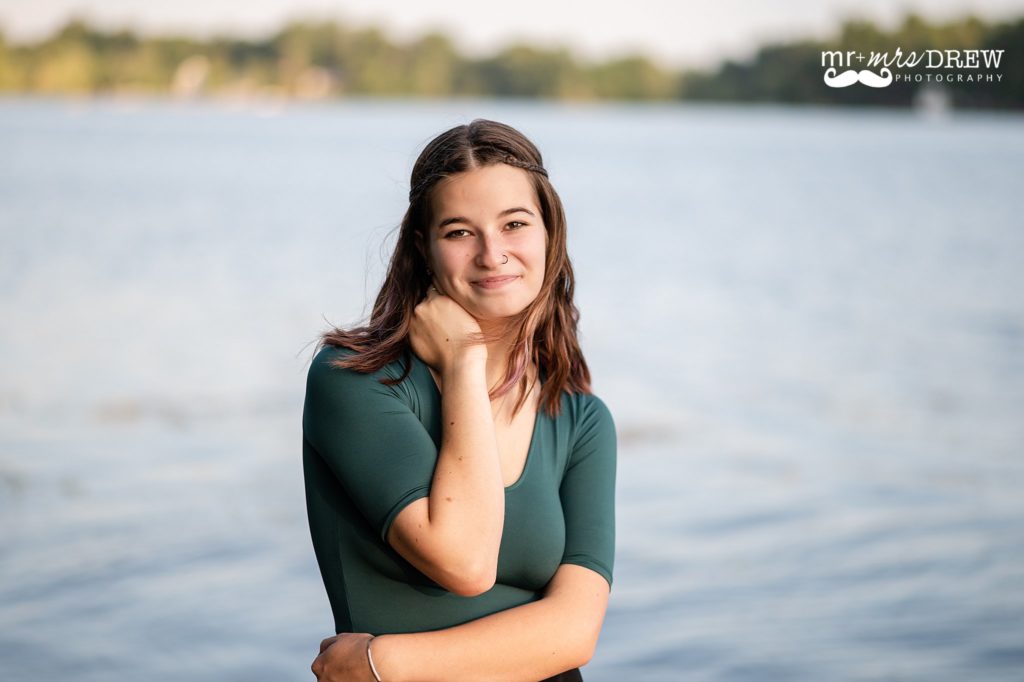 Senior Photos at Forge Pond Westford MA. Senior posed in front of the water holding onto her neck wearing a green shirt and smiling. 
